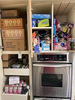 Shelving unit holding multiple boxes of LED light bulbs, miscellaneous packing bags, disposable gloves, and spray cans above and next to a stainless steel oven.
