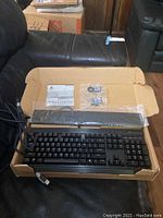 Corsair K70 LUX RGB keyboard in open cardboard box with attached USB cable, wrist rest, replacement keycaps, and manual.