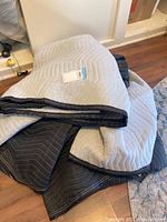 Four large double-sided furniture blankets shown stacked and spread on hardwood floor near a white wall and rug. Blankets feature a light gray side and a dark gray side with quilting stitched in zigzag patterns and black edging.