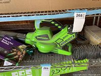 Greenworks cordless leaf blower with a box showing an Orbit brass 4-port manifold and a green garden hose spray nozzle on a metal shelf.