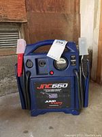 Front view of JNC 660 jump starter device showing brand and model label, analog gauge, power button, and jumper cables attached.