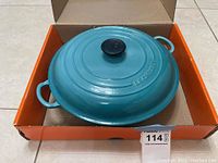 Top view of covered blue Le Creuset pan inside an orange cardboard box showing the lid with a black knob and two side handles.