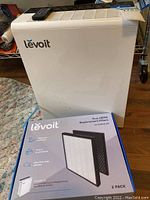 White Levoit air purifier and box of filters on a wooden floor with remote control on top of purifier.