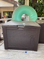 Dark brown outdoor resin storage box with wood-effect lid on a wooden porch. The box has textured sides resembling rattan and a front handle.