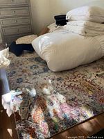 Floral multicolored area rug laid on a wooden floor with bedding and miscellaneous items on top.