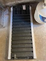 Photo of the full motorbike ramp showing grooved traction surface and metal edges, lying on concrete floor beside propane tank.