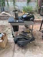 Full view of Weber charcoal BBQ on metal stand with chimney charcoal starter on side and BBQ cover below.