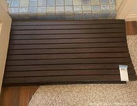 Rectangular dark brown wood slat bathroom mat positioned on floor in front of mosaic tiled threshold, showing full mat and some surrounding area.