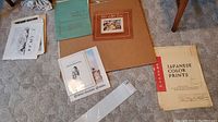 Loose prints laid out on carpet including a Japanese color print, a mounted print folder, and other loose prints with some visible wear.