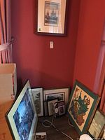 Collection of framed prints and artwork leaning against wall in corner of room. Includes framed prints and a batik recording photo frame with key card.