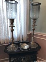 Two tall hurricane candle holders with etched glass shades on ornate brass bases next to two metal bowls on a wooden surface