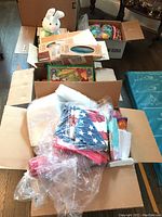 Boxes filled with various holiday party supplies and decorations including patriotic napkins and cups, plastic plates, and Easter eggs.