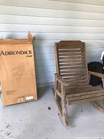 Weathered wooden rocking chair with slatted design alongside a new boxed Adirondack chair in original packaging, positioned on a porch with light blue siding background.