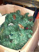 Box containing artificial green Christmas garland decorated with pine cones and red berries. Visible packing and decor context.