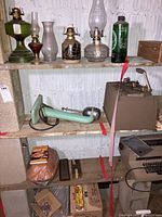 View of shelves showing five oil lamps, green handheld blender, Oster fondue set, Sterno canned heat, Romertopf cookware and cookbook.