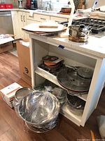 Photo showing white cabinet with multiple aluminum and stainless steel pots and pans inside, also cutting boards on top of the cabinet