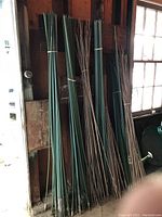 Several bundles of green-coated metal garden stakes and uncoated rusty metal stakes, all bundled together leaning against a wall in a shed or garage.
