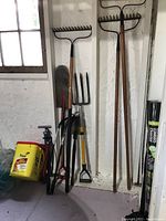 Various garden tools including rakes, shovels, pitchfork, and bicycle pumps leaning against a white textured wall next to a window.