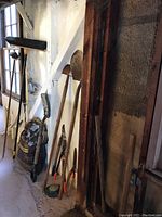 Photo of garden tools including shovels, loppers, shop broom, mulch bag, sledgehammer, and pickaxe leaning against a white wall indoors showing weathered condition.