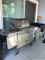 Front side view of Weber gas grill showing stainless steel surfaces, rusted legs, side shelves, and storage cabinet with double doors.