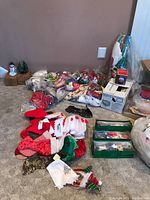 Wide view of assorted Christmas decorations including tablecloths, cookie cutters, ceramic items, gift wrap, and other packaging supplies loosely arranged on carpet against a wall.