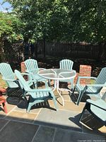 Outdoor patio setting showing round glass top table with white metal frame and six green plastic Adirondack-style chairs arranged around it on stone patio.