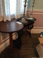 Two wooden side tables against wall, one round pedestal and one rectangular with ornate legs, showing surface scratches and wear.