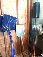 Two large blue metal snow shovels and one smaller silver shovel leaned against a wooden wall next to a metal bird feeder hook with cylindrical black container.