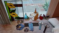 Shows all stained glass pieces: one large rectangular panel with two large yellow flowers and 11 smaller assorted stained glass items including a unicorn, dragonfly, sailboat, apple, flowers, butterflies, and other shapes.