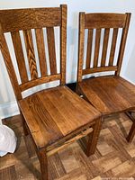 Pair of vintage oak straight back side chairs standing against white wall, showing full view of front and sides with visible scratches on seats.