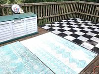 Outdoor storage bin on wooden deck with two identical reversible outdoor mats laid on deck, turquoise and white geometric patterned sides visible.