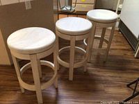 Three matching solid wood counter height stools with round seats and swivel mechanism on a wood floor.