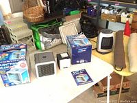 Lot contents on table including two boxed Arctic Air Pro small evaporative coolers, two coolers out of box, small Holmes space heater, 2 door insulators rolled up.