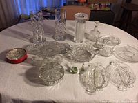 Overview photo showing all cut glass items including vases, serving trays, decanter, lidded containers, and decorative birds on a white tablecloth.