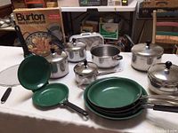 Overview of Farberware stainless steel pots with black knob lids, green Orgreenic frying pans stacked, and box for Burton Maxi Stovetop Grill on table