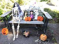 Full view of skeleton, witches, pumpkins, spider web and masks arranged on and near a wooden bench outdoors