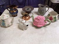 Full view of seven tea pots and pitchers including cobalt blue and copper lusterware pitcher and six porcelain pots with floral and unique designs.