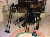 Photo showing two black canes with quad rubber bases, a metal safe step stool with black step, three black walking sticks with rubber grips, and part of a black rollator walker with seat and storage bag.