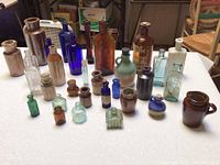 Photo showing entire assortment of vintage bottles and ceramic jars with varied colors, sizes, and shapes on white tablecloth.