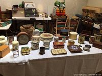 Photo showing vintage tins of various shapes and sizes arranged on a table with a wicker basket and other vintage items in the background.