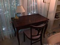 Full view of the mahogany writing desk with the brass table lamp on top and the antique chair pushed in. The desk top has a polished finish with a decorative border. The table lamp has a white pleated lampshade.