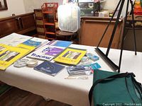 Full spread of watercolor paper pads, tracing paper, artist brushes, palettes, and carrying bag laid out on white tablecloth with a wooden floor and furniture background.