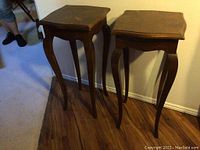 Two Italian wood plant stands with curved legs and scalloped square tops, positioned side by side on wood flooring, showing wear on tops.