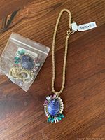 Gold-tone necklace with large oval purple stone and colorful accent stones, displayed with matching bracelet in plastic bag