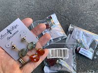 Close-up of a pair of dangling earrings featuring green and orange stones held in hand with packaging in the background.