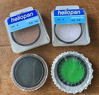 Four 55mm SLR lens filters shown on wooden surface, two in original Heliopan packaging labeled KR 3 ES 56 and KR 6 ES 55, two wrapped in clear plastic coverings.