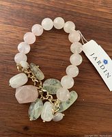 Close-up photo of a single bracelet with large round pale pink beads and multiple semi-precious stone charms attached to a gold-tone chain. Bracelet is tagged with Jardin brand label.