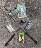 Photo of marshmallow roasting forks crossed on concrete with utility tool, bear bell, stretch cords, travel containers and bungee cords visible on the ground.