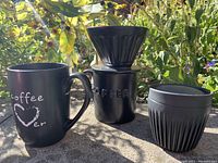 Outdoor photo of three black coffee mugs including a large mug with white text, a coffee drip cone on a matching mug, and a travel mug