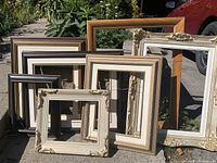 Seven empty picture frames arranged outdoors showing variety of colors, sizes, and styles including dark wood, light wood, and ornate cream frames